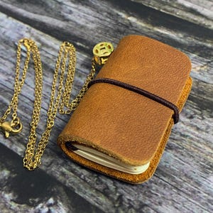 Peut inclure: Un carnet en cuir marron avec une chaîne dorée et un petit charme doré. Le carnet est fermé et possède un élastique noir autour.
