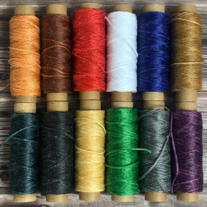 May include: A collection of twelve spools of waxed thread in various colors, including orange, brown, red, white, blue, and gold. The spools are arranged in two rows, with each spool tightly wound and ready for use. The background is a wooden surface.