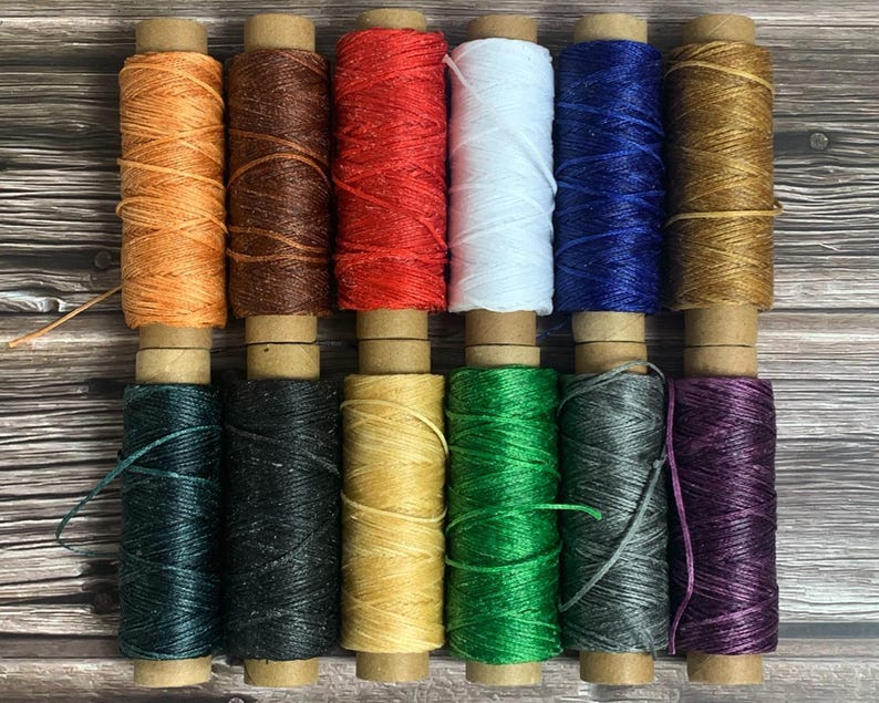 May include: A collection of twelve spools of waxed thread in various colors, including orange, brown, red, white, blue, and gold. The spools are arranged in two rows, with each spool tightly wound and ready for use. The background is a wooden surface.