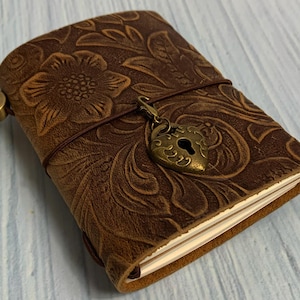 May include: A brown leather journal with a floral embossed design and a heart-shaped lock charm. The journal is closed and has a brown string wrapped around it.
