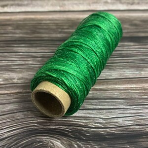 May include: A spool of bright green thread, tightly wound around a cardboard core, rests on a wooden surface. The thread has a slight sheen, and the color is a vibrant, saturated green. The wooden surface has a natural, weathered appearance.