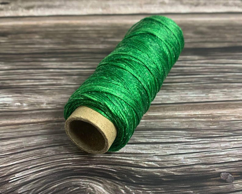May include: A spool of bright green thread, tightly wound around a cardboard core, rests on a wooden surface. The thread has a slight sheen, and the color is a vibrant, saturated green. The wooden surface has a natural, weathered appearance.