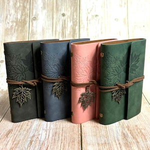 Op de afbeelding: Vier leren journals in grijs, blauw, roze en groen. Elk journal heeft een reliëf bladontwerp en is vastgemaakt met een bruin leren koord en een metalen bladbedel. De journals worden gepresenteerd op een houten oppervlak. De journals zijn ongeveer 15 cm hoog.