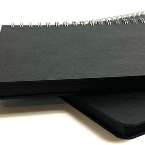 Puede incluir: Dos cuadernos negros de espiral apilados uno encima del otro. Los cuadernos est&aacute;n cerrados y la encuadernaci&oacute;n es visible.