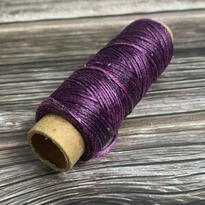 May include: A spool of purple waxed thread. The thread is tightly wound around a cardboard core. The color is a deep, rich purple with a slight sheen. This thread is likely used for crafting or jewelry making.