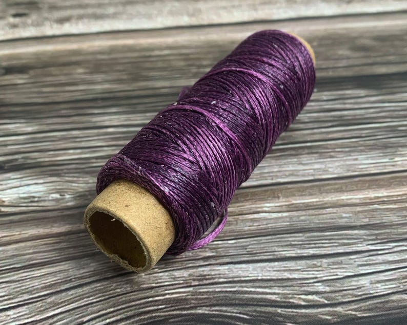 May include: A spool of purple waxed thread. The thread is tightly wound around a cardboard core. The color is a deep, rich purple with a slight sheen. This thread is likely used for crafting or jewelry making.