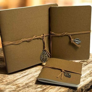 May include: Three brown kraft paper journals of varying sizes, bound with twine and adorned with metal leaf charms. The journals are arranged on a rustic wooden surface, creating a natural and earthy aesthetic.