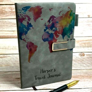 Könnte beinhalten: Graues Reisetagebuch mit farbenfrohem Weltkarten-Design. Das Tagebuch hat einen goldenen Verschluss und den Text "Harper's Travel Journal" auf dem Einband. Ein goldener und schwarzer Stift liegt auf der Oberfläche.