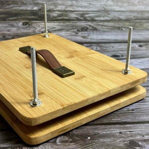 Può includere: Una pressa per piante in legno con manico in pelle marrone e ferramenta in metallo. La pressa è realizzata in legno chiaro e tenuta insieme da quattro lunghi bulloni metallici. È progettata per pressare fiori e piante.