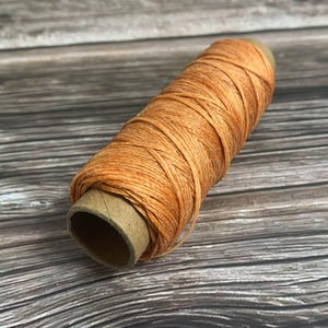 May include: A spool of orange-colored thread, tightly wound around a cardboard core. The thread has a slightly textured appearance, and the spool is positioned diagonally against a wooden background.