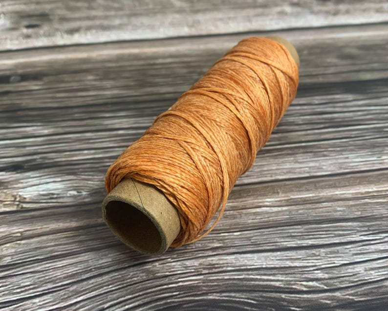May include: A spool of orange-colored thread, tightly wound around a cardboard core. The thread has a slightly textured appearance, and the spool is positioned diagonally against a wooden background.