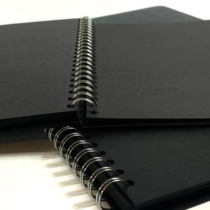 Puede incluir: Un cuaderno negro de espiral con una cubierta negra. El cuaderno est&aacute; cerrado y la encuadernaci&oacute;n es visible.