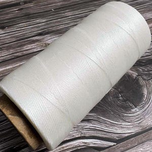 May include: A large spool of white thread, tightly wound, with a cardboard core. The thread has a slightly textured appearance. The spool is resting on a dark wood surface.