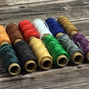 May include: A selection of colourful waxed linen thread spools in shades like orange, brown, red, white, blue, and green. The spools are displayed on a wooden surface, highlighting the diverse colours of the thread.