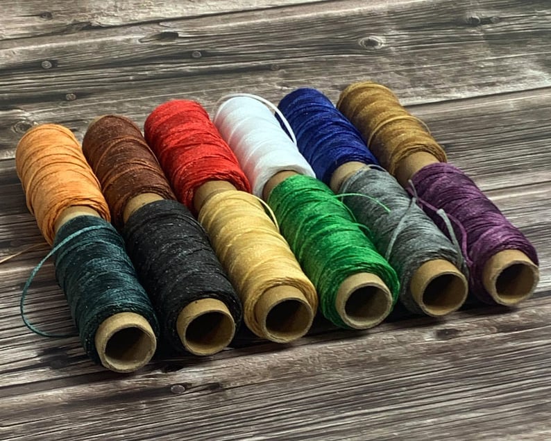 May include: A collection of colorful waxed linen thread spools in various colors, including orange, brown, red, white, blue, and green. The spools are arranged on a wooden surface, showcasing the vibrant hues of the thread.