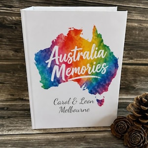 May include: A white photo album featuring a rainbow-coloured watercolour map of Australia. The words "Australia Memories" are written across the map. The names "Carol & Leon" and "Melbourne" are printed below.