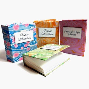 Peut inclure: Quatre carnets de voyage décoratifs avec des couvertures marbrées. Les carnets sont étiquetés "Venice Memories", "Paris Memories" et "Anna & Steph Travels". Un carnet est ouvert, affichant des pages vierges. Les couvertures présentent des motifs marbrés bleus, oranges, roses et verts.