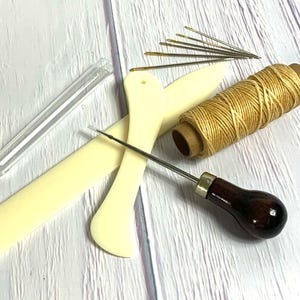 May include: A set of leatherworking tools including a stitching awl, two leather stitching tools, a spool of waxed thread, and a needle threader.
