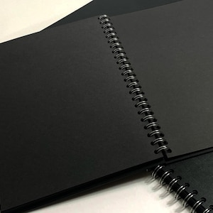 Puede incluir: Un cuaderno negro de espiral con una cubierta negra. El cuaderno est&aacute; abierto para revelar p&aacute;ginas en blanco.