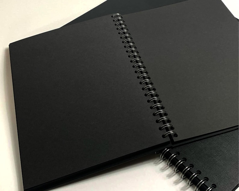 Puede incluir: Un cuaderno negro de espiral con una cubierta negra. El cuaderno est&aacute; abierto para revelar p&aacute;ginas en blanco.