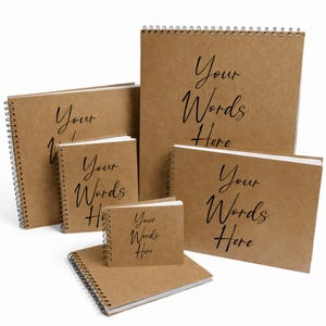 May include: A collection of spiral-bound notebooks in various sizes. The covers are a natural brown color, and each notebook features the text "Your Words Here" in a black script font. The notebooks are arranged in a group, showcasing their different dimensions.