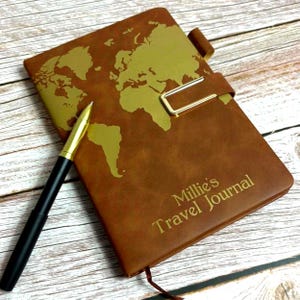 Könnte beinhalten: Braunes Leder-Reisetagebuch mit goldener Weltkarte und goldenem Verschluss. Das Tagebuch trägt den Text "Millie's Travel Journal". Ein schwarzer und goldener Stift liegt auf dem Tagebuch.