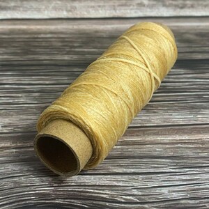 May include: A spool of light tan thread, tightly wound around a cardboard core. The thread has a slightly textured appearance and is crisscrossed in a few places. The background is a wooden surface.