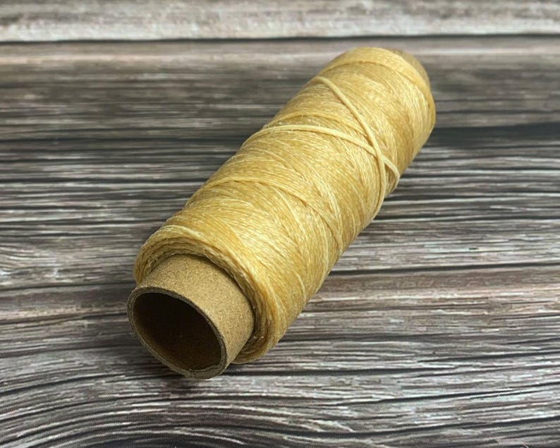 May include: A spool of light tan thread, tightly wound around a cardboard core. The thread has a slightly textured appearance and is crisscrossed in a few places. The background is a wooden surface.