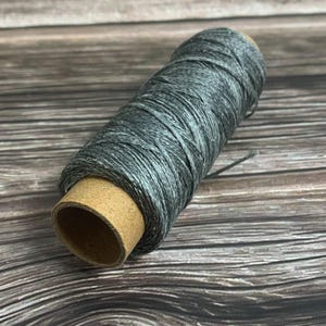 May include: A spool of gray thread, tightly wound around a cardboard core. The thread has a subtle, variegated color, with shades of gray and white. The spool is positioned diagonally, showcasing the texture of the thread.