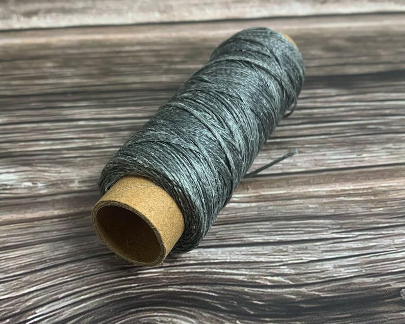 May include: A spool of gray thread, tightly wound around a cardboard core. The thread has a subtle, variegated color, with shades of gray and white. The spool is positioned diagonally, showcasing the texture of the thread.