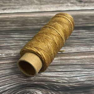 May include: A spool of golden-brown waxed thread. The thread is tightly wound around a cardboard tube. The spool is positioned diagonally, showcasing the texture and color of the thread. Ideal for crafting and sewing projects.