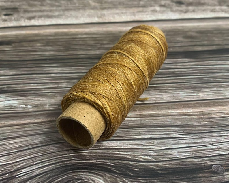May include: A spool of golden-brown waxed thread. The thread is tightly wound around a cardboard tube. The spool is positioned diagonally, showcasing the texture and color of the thread. Ideal for crafting and sewing projects.