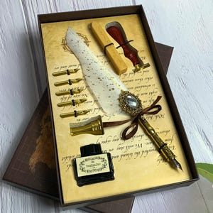 May include: A vintage calligraphy set in a decorative box. The set includes a quill pen with a jeweled embellishment, ink bottle, multiple nibs, sealing wax, and a wax stamp. The box has a parchment-like background with script writing.