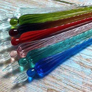 Peut inclure: Une collection de stylos à plume en verre coloré. Les stylos sont disposés en diagonale, présentant une variété de couleurs, dont le transparent, le vert, le marron, le rouge, le rose, le turquoise et le bleu. Chaque stylo a une plume détaillée et une extrémité arrondie.