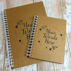 Può includere: Due quaderni rilegati a spirale con copertine in carta kraft. Il quaderno più grande è aperto, rivelando la rilegatura a spirale bianca. Il quaderno più piccolo è chiuso. Entrambi i quaderni presentano il testo "Your Words Here" in corsivo nero, con illustrazioni di cuori.