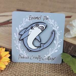 May include: Enamel pin featuring a cartoon shark with a surprised expression. The shark is light blue with a white belly and black outlines. The pin is on a light blue card with the text "Enamel Pin" and "Pinku's Crafty Cottage".