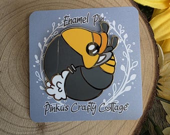 May include: Enamel pin featuring a stylized insect design with yellow, black, and white colors. The pin is on a gray card with the text "Enamel Pin" and "Pinku's Crafty Cottage". The pin is set against a wooden background.