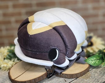 May include: A plush backpack shaped like a pill bug, featuring a white, brown, and yellow color scheme. The backpack has a large, round body with a detailed eye and a gray strap. It is placed on a wooden slice.