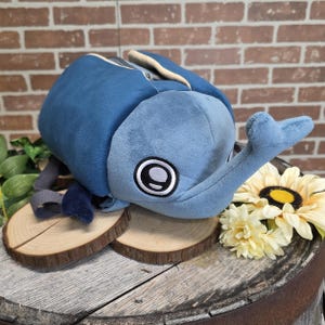 May include: A plush blue beetle-shaped backpack with a zippered top and a light blue head and antennae. The backpack has a large eye design on the side and is displayed on wooden slices with a brick background.