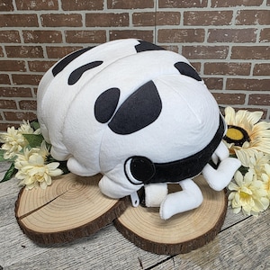 May include: A plush toy shaped like a pill bug, featuring a white body with black spots. The toy has a black band around its head and small white appendages. It rests on two wooden slices, with flowers in the background.