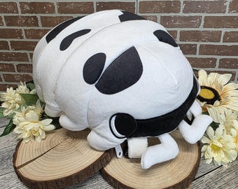 May include: A plush toy shaped like a pill bug, featuring a white body with black spots. The toy has a black band around its head and small white appendages. It rests on two wooden slices, with flowers in the background.