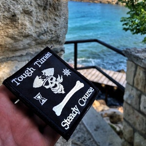 以下が含まれることがあります： 白いドクロと交差した骨の刺繍が施された黒いパッチ。パッチには「Tough Times Steady Course」という文字が書かれています。