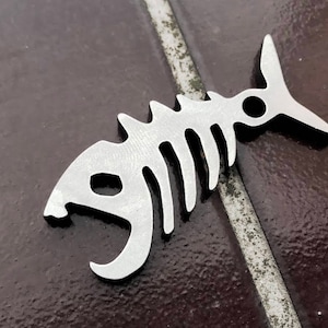 May include: A silver metal fish skeleton bottle opener. The opener is shaped like a fish with a detailed skeleton design. The bottle opener has a small hole at the top for attaching a keychain.