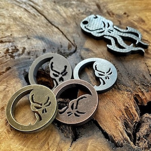 Peut inclure: Un ensemble de jetons métalliques et un petit porte-bonheur en forme de poulpe. Les jetons sont circulaires avec un motif de crâne découpé au centre. Les jetons ont une finition bronze, cuivre et argent. Le porte-bonheur poulpe est argenté.