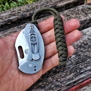 Könnte beinhalten: Ein silbernes Taschenmesser mit einem grünen Paracord-Lanyard. Das Messer hat eine kleine Klinge und einen Sicherheitsverschluss. Das Messer ist geschlossen und das Lanyard ist in einem Knoten gebunden.