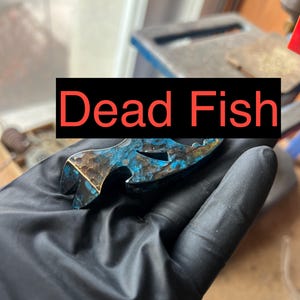 Könnte beinhalten: Ein handgefertigter Flaschenöffner mit blauem und bronzefarbenem Finish. Der Text "Dead Fish" wird in einem schwarzen Rechteck angezeigt. Der Öffner wird in einer schwarz behandschuhten Hand gehalten.