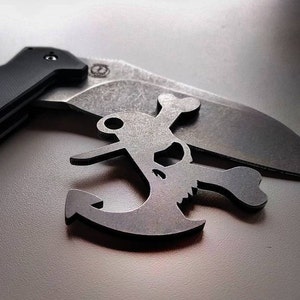 Pode incluir: Um chaveiro de metal com um design de caveira e ossos cruzados, em forma de âncora. O chaveiro é de cor prateada e tem um acabamento desgastado.