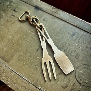 Op de afbeelding: Een bronzen sleutelhanger met een vork- en spatelbedel. De bedels zijn verbonden met een sluiting en een ring. De sleutelhanger rust op een verweerd, olijfgroen oppervlak, wat wijst op een rustieke of vintage esthetiek.
