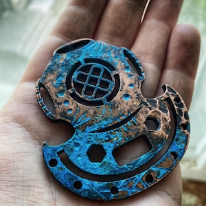 Peut inclure: Un pendentif en métal cuivre et bleu en forme de casque de plongée avec un motif de nœud celtique au centre. Le pendentif a un aspect vieilli et usé.