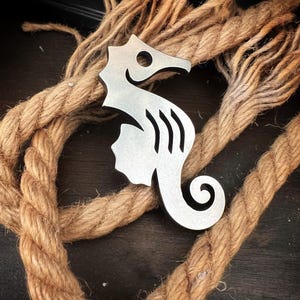 May include: A silver metal seahorse keychain with a detailed design. The keychain is lying on a brown rope.